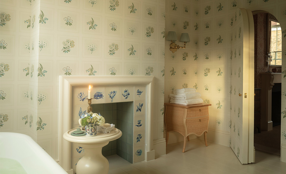 Etoile Blue fabric in a bathroom with a blue and white wallpaper and tiled fireplace and a white bathtub, table and candle and small rattan chest