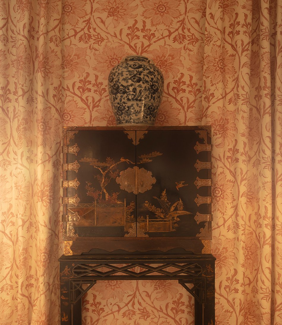 Palampore Blossom Indian Pink wallpaper in a bedroom an antique trunk and vase 