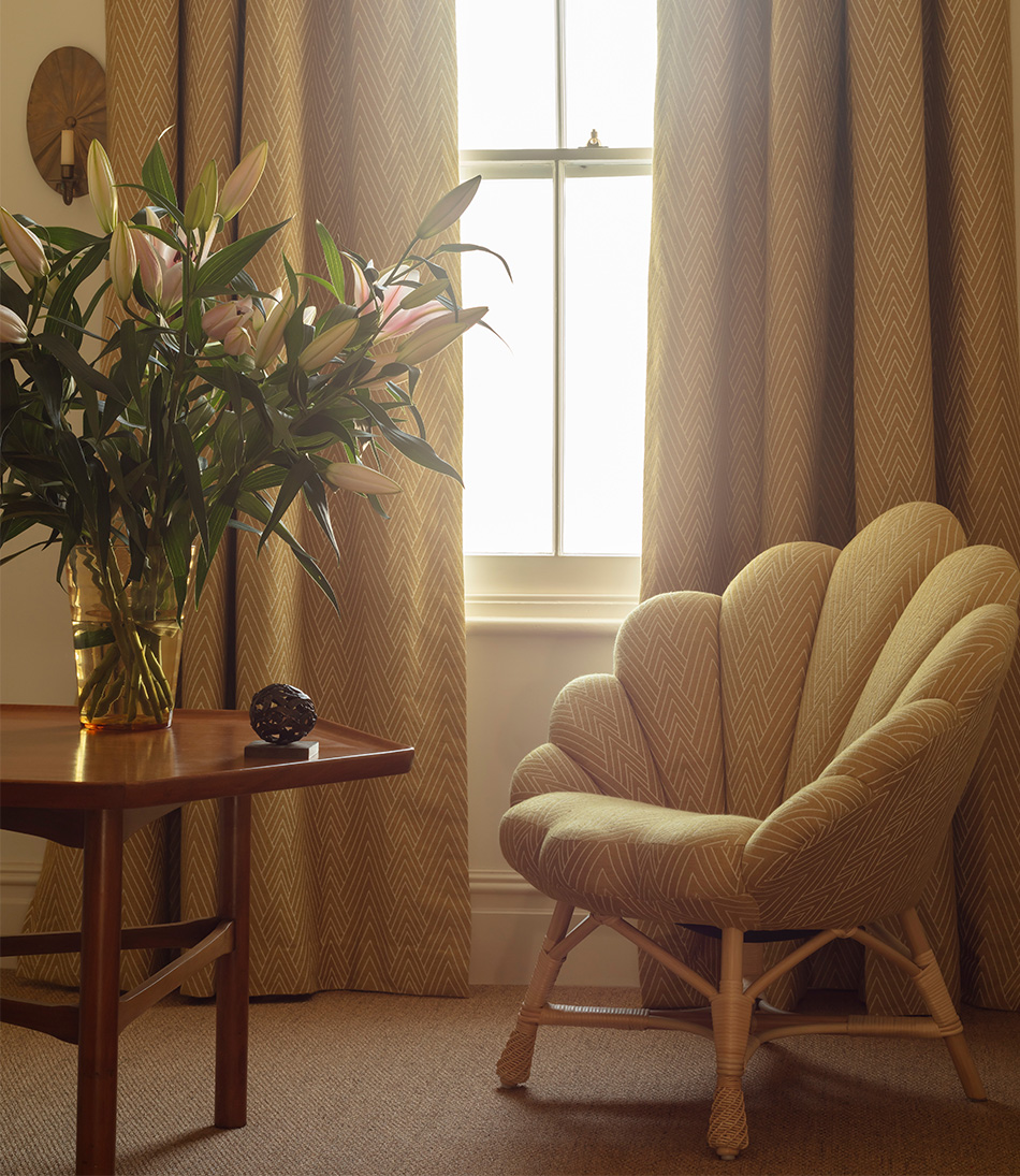 Philae Old Gold upholstered on a chair and curtains with a low wooden table and flowers