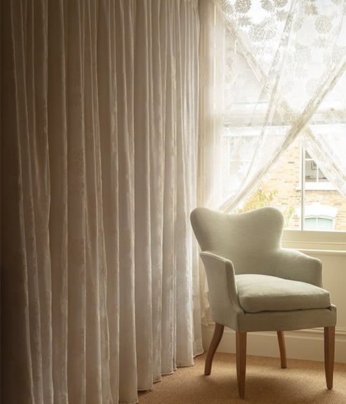 The Gigi chair in a pale blue fabric and shown with a lace curtain 